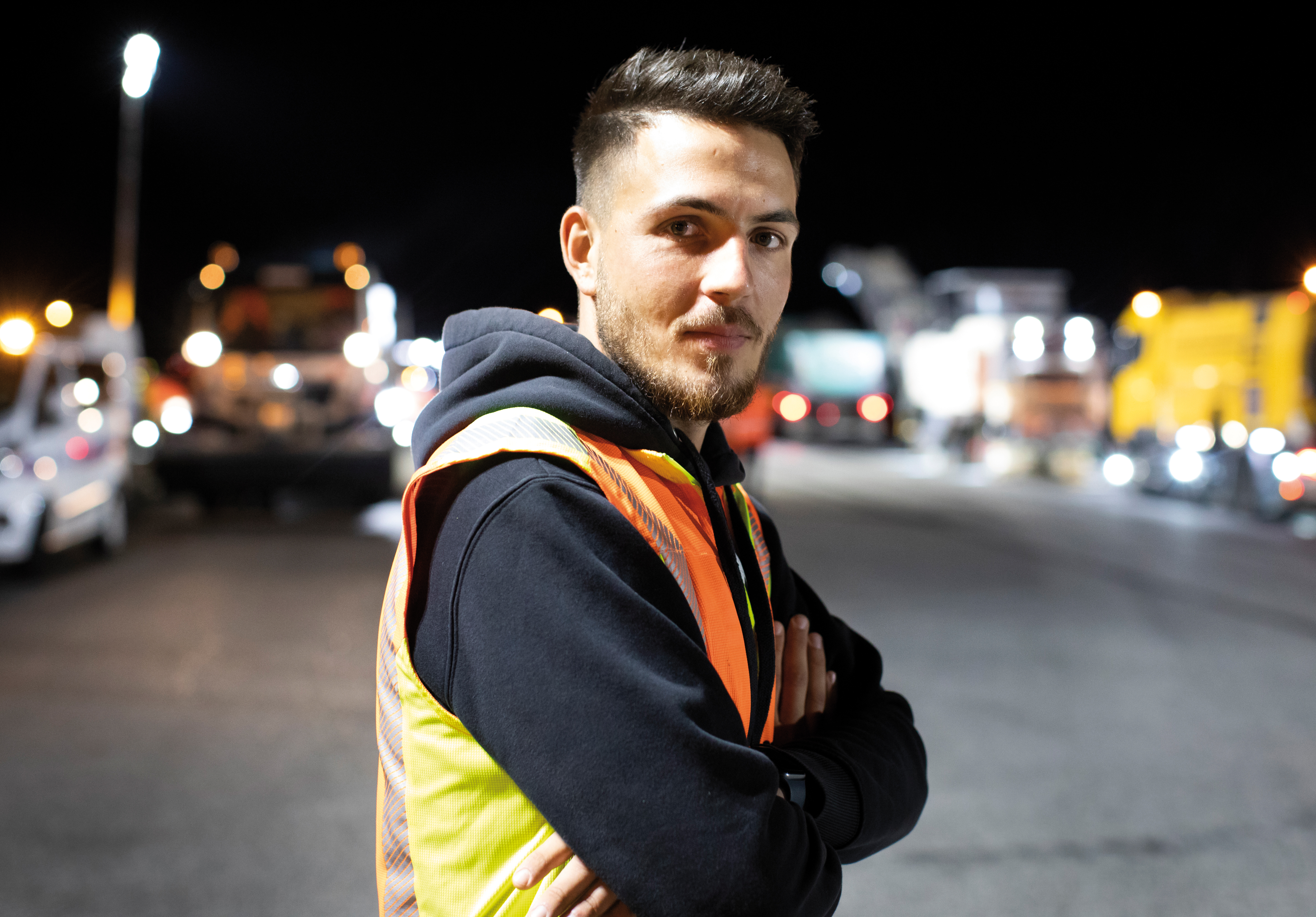 Seitlich stehender Mann mit verschränkten Armen, trägt eine Warnschutzweste über einem schwarzen Kapuzenpullover.