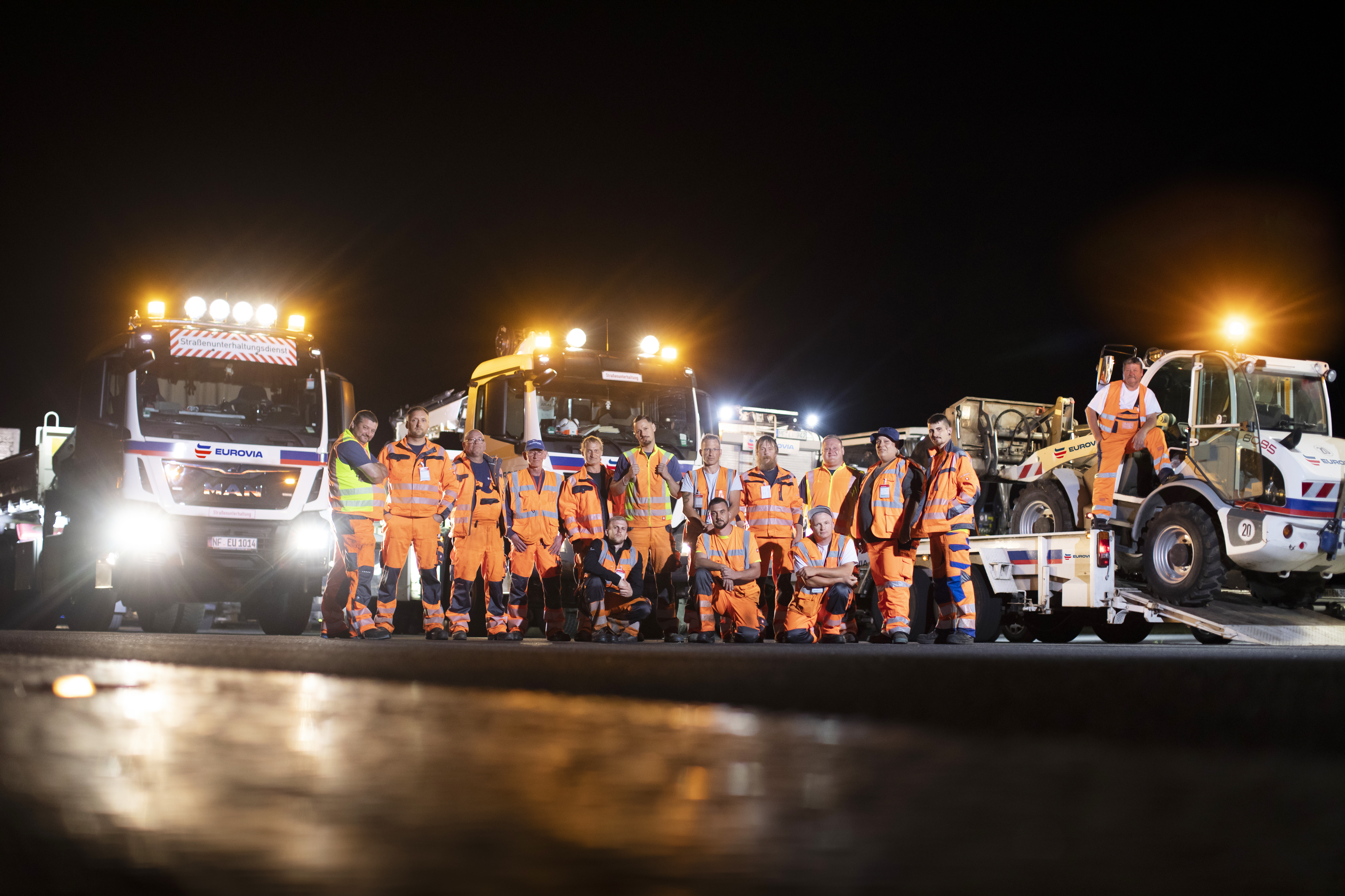EUROVIA-Team mit 15 Personen steht nachts vor EUROVIA-LKWs und Maschinen