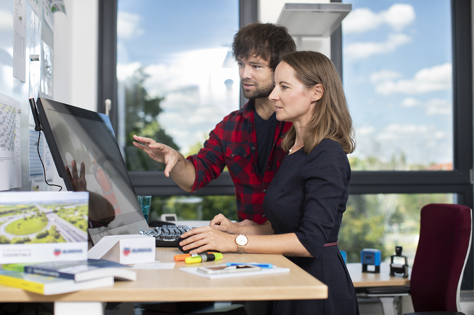 Mann und Frau stehen an einem Stehschreibtisch, Mann erklärt der Frau etwas am Monitor.