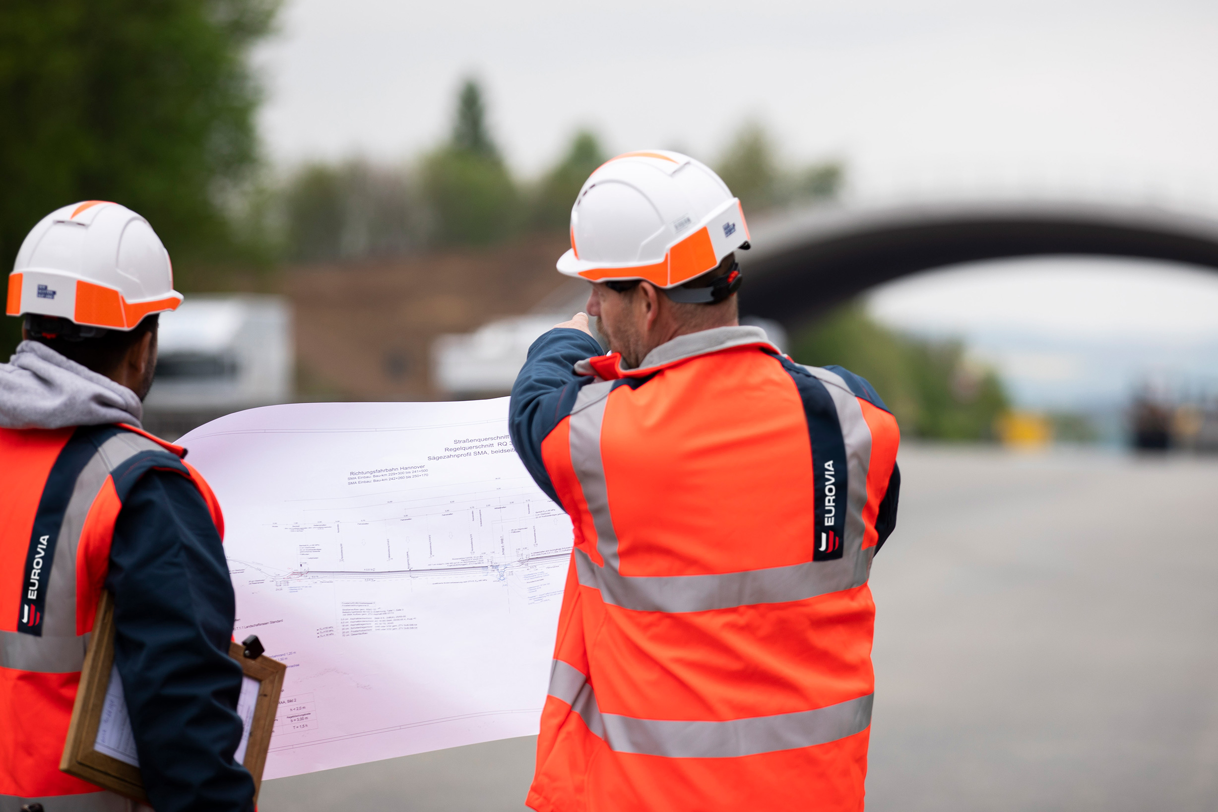 Zwei Mitarbeitende, die mit dem Rücken zur Kamera stehen und einen Plan in der Hand halten.