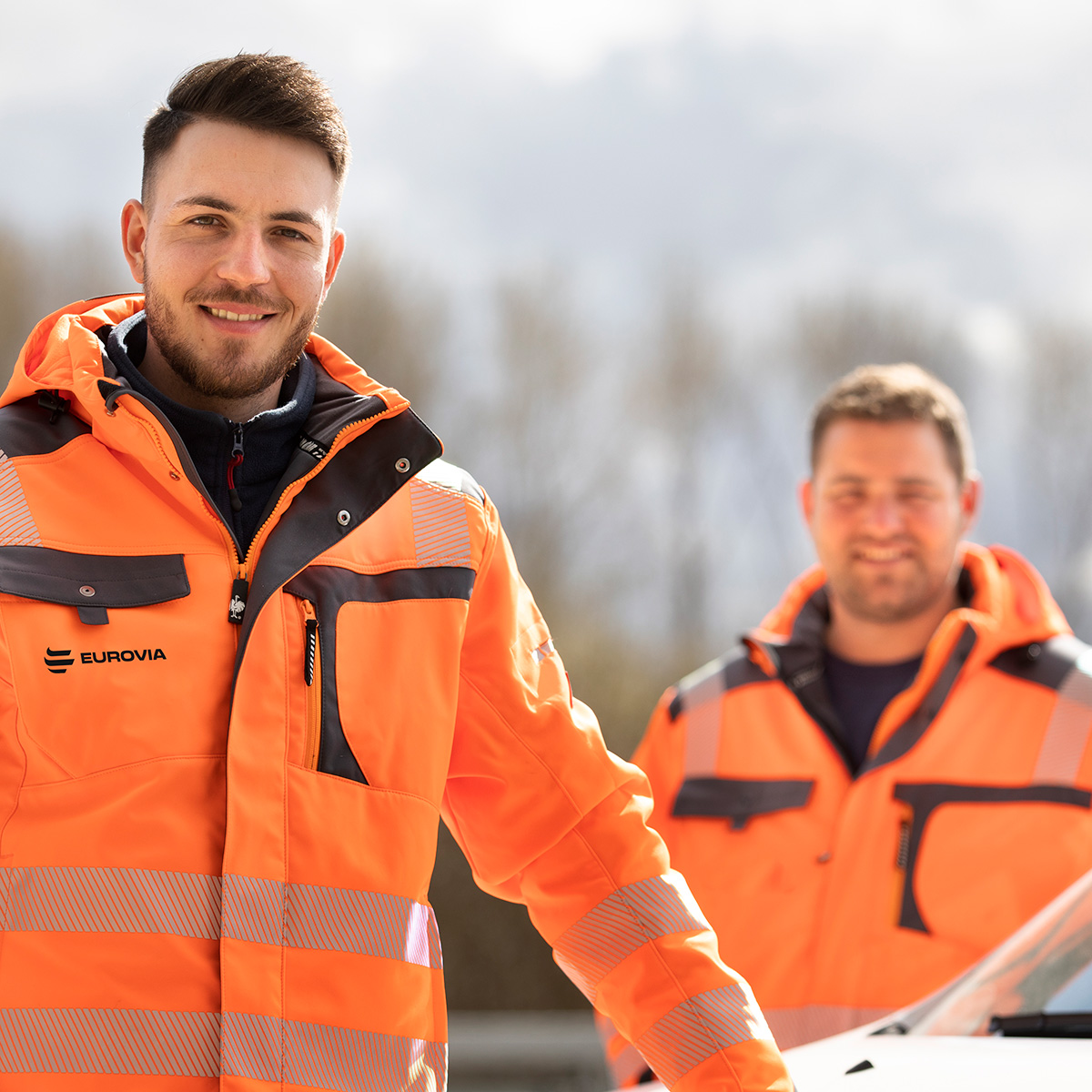 Zwei Arbeiter in Warnschutzjacken, vorderer Mann freundlich im Fokus.