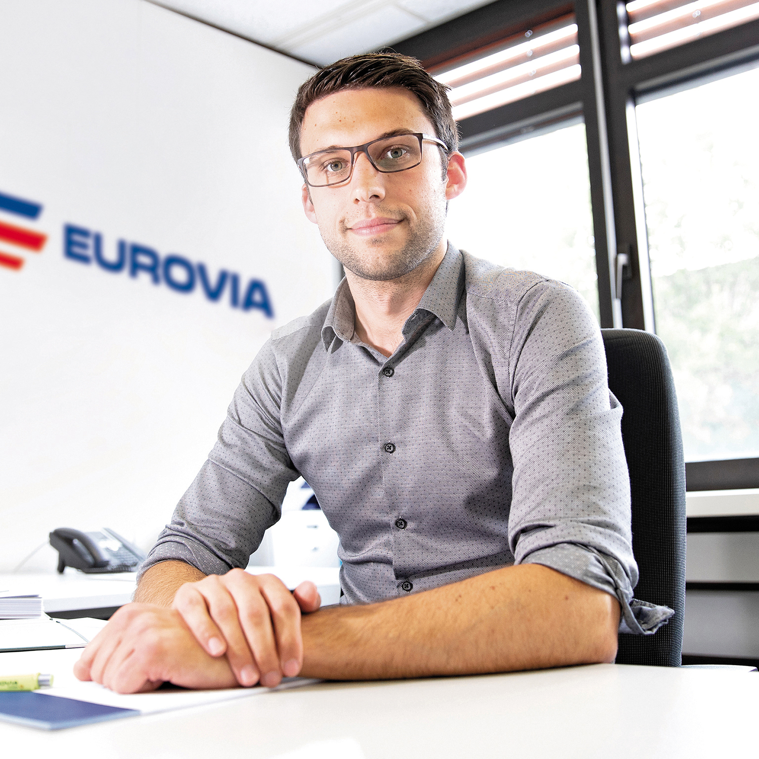 Mann im grauen Hemd sitzt am Schreibtisch, weiße Wand mit EUROVIA-Logo im Hintergrund.