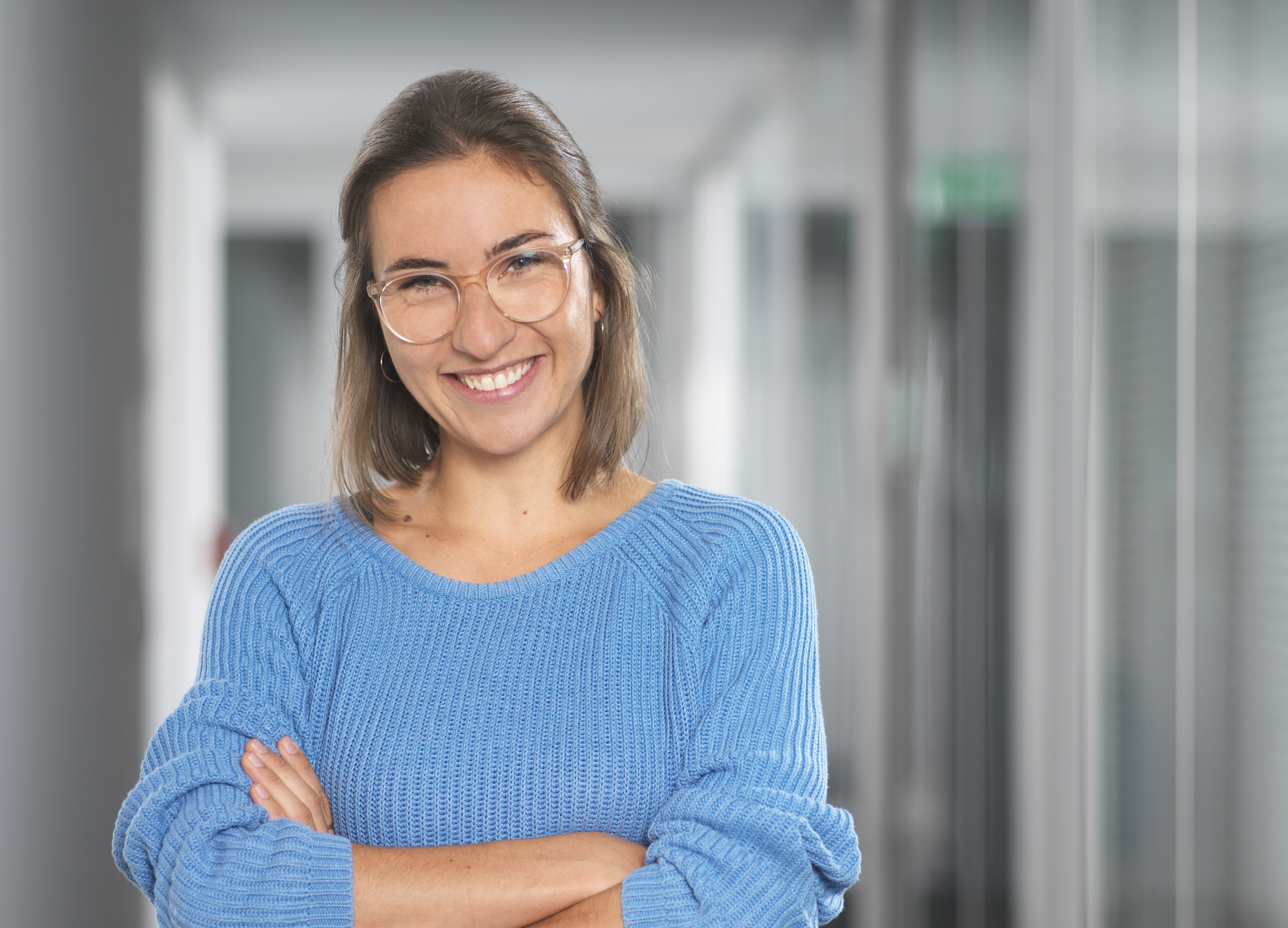 Frau, mit blauem Pullover und verschränkten Armen lächelt frontal.
